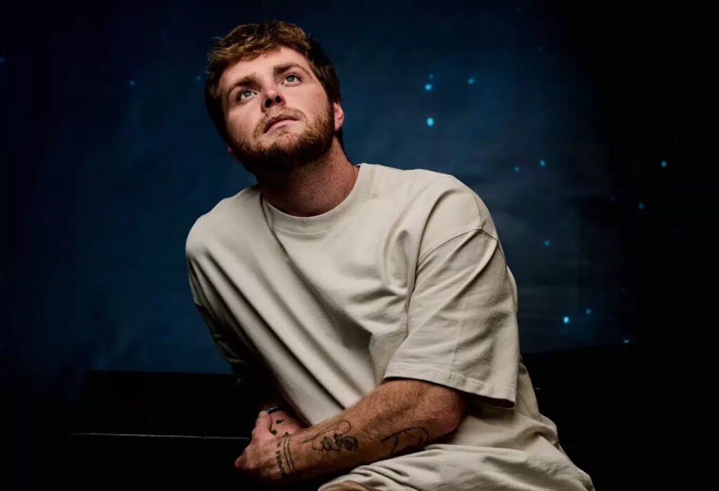 Alex Warren sits in front of a dark navy back drop looking up to the sky