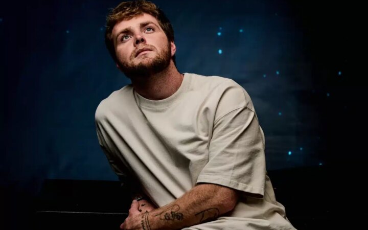 Alex Warren sits in front of a dark navy back drop looking up to the sky