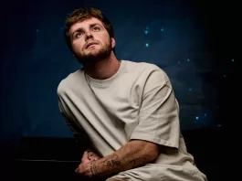 Alex Warren sits in front of a dark navy back drop looking up to the sky