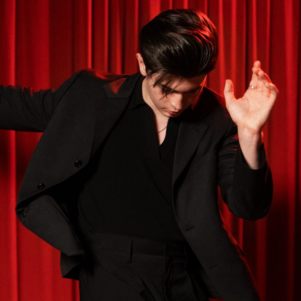 Elliot James Reay in a black suit in front of red drapes